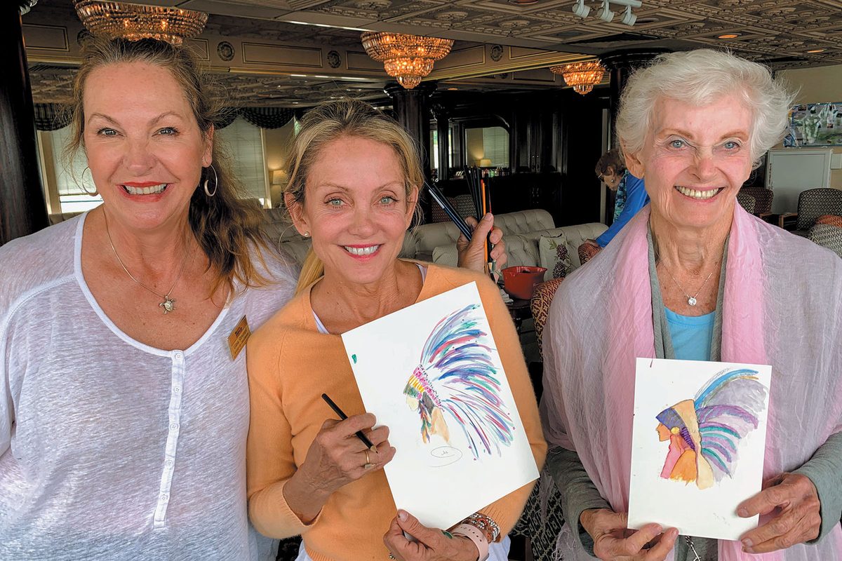 Three smiling women hold up their colorful watercolor paintings after an onboard art class