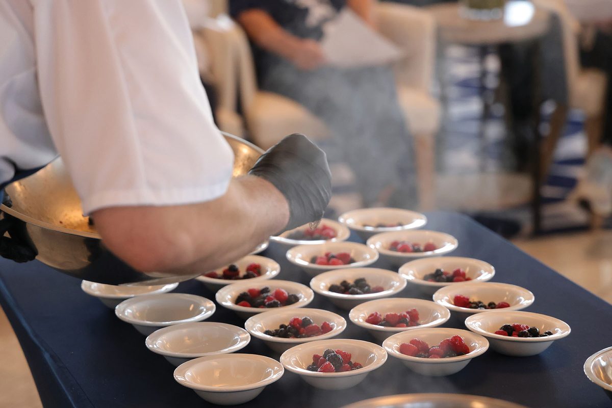 A chef spoons fresh mixed berries into rows of small white bowls during a cooking demonstration