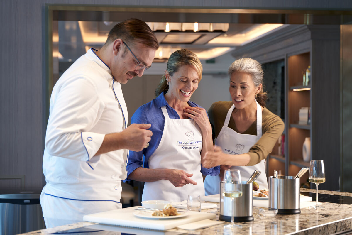 A chef guides two smiling guests in aprons during a hands-on cooking class at the Oceania Cruises Culinary Center