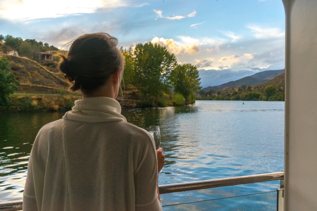 A traveler holds a glass of wine at the railing while taking in a serene river and hillside vineyard view
